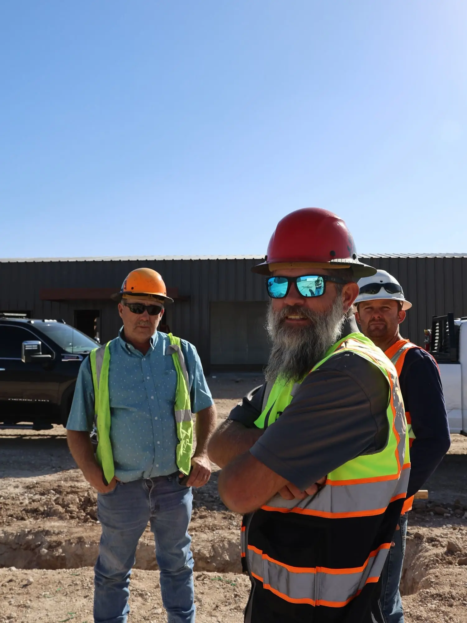 Three crew members in hard hats and safety vests on a job site in bright sun
