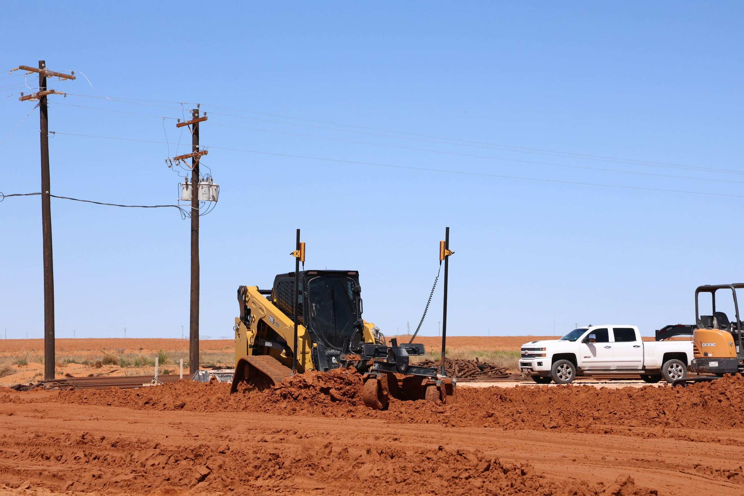 Skid steer grading a red dirt pad beside a pickup and a mini excavator under power lines
